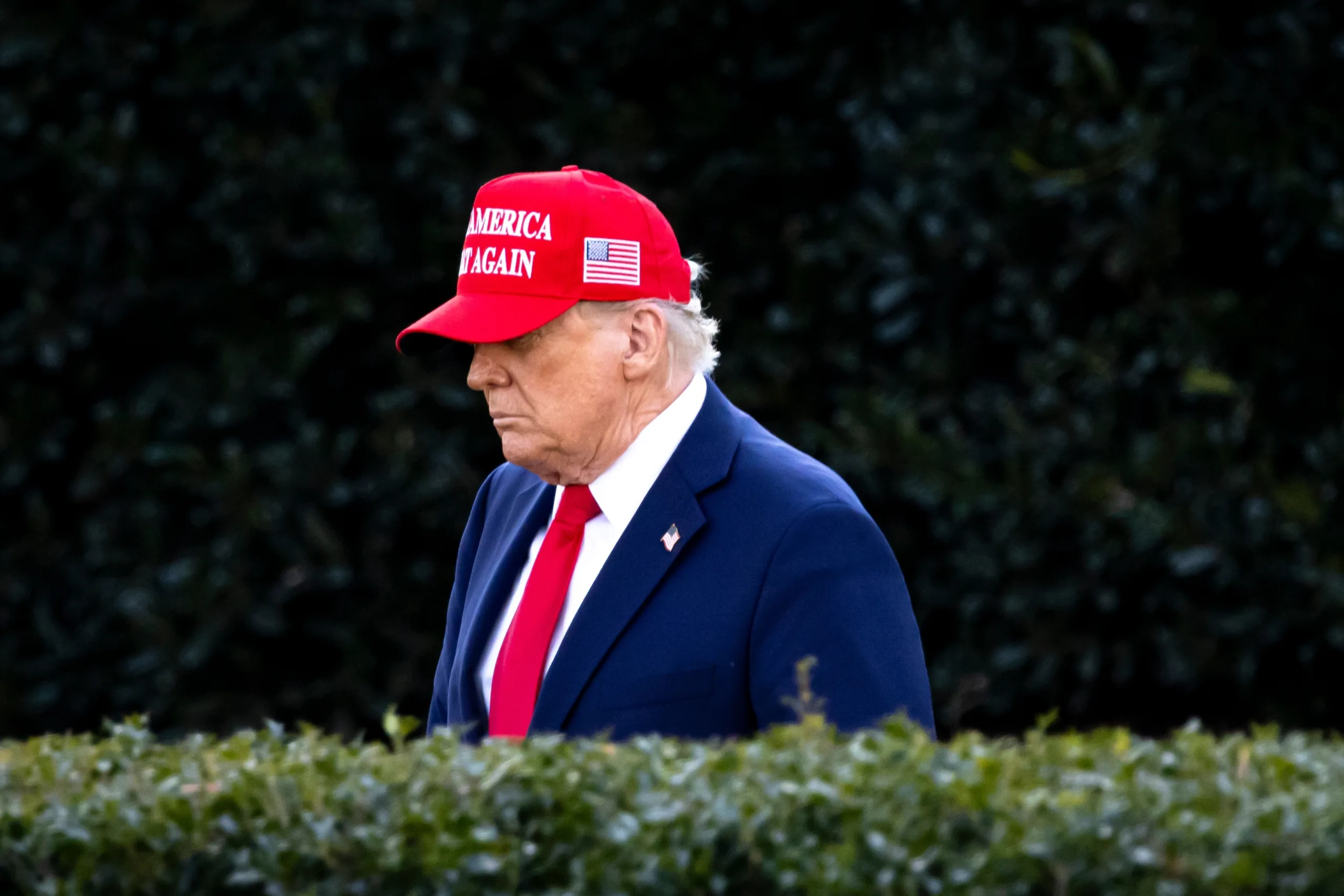 jvhlf يرتدي بدلة رسمية وقبعة حمراء مكتوب عليها "اجعل أمريكا عظيمة مرة أخرى" يقف في الهواء الطلق