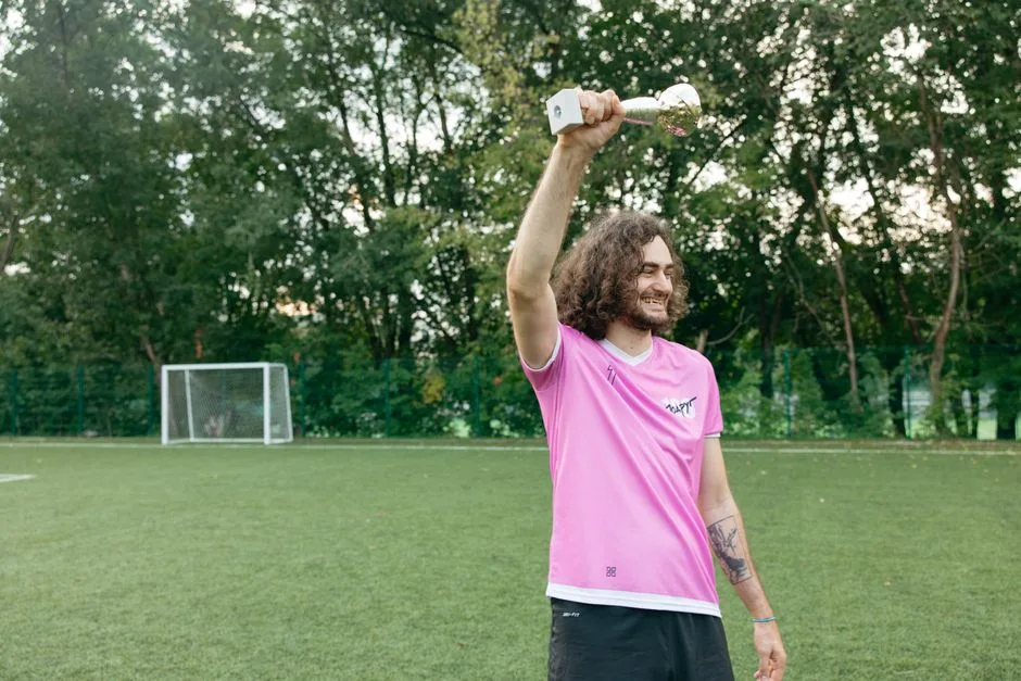 Smiling soccer player with curly hair celebrates a victory holding a trophy on an outdoor field.